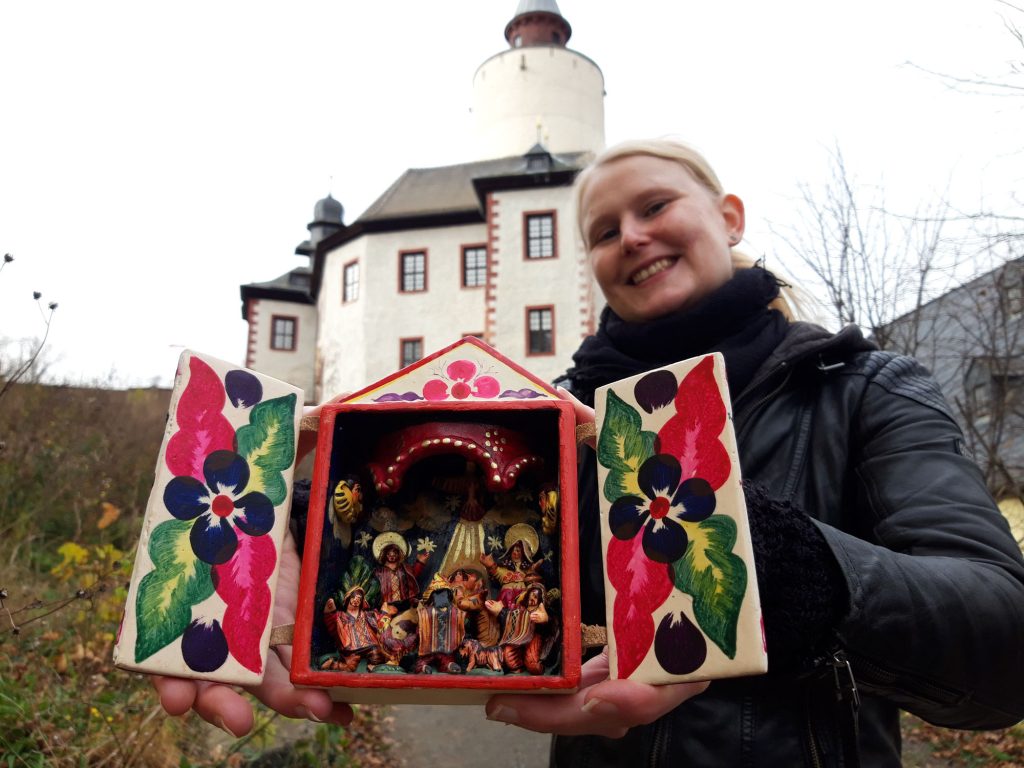 Junge Frau hält eine bunte südamerikanische Krippe in der Hand, im Hintergrund die Burg Posterstein