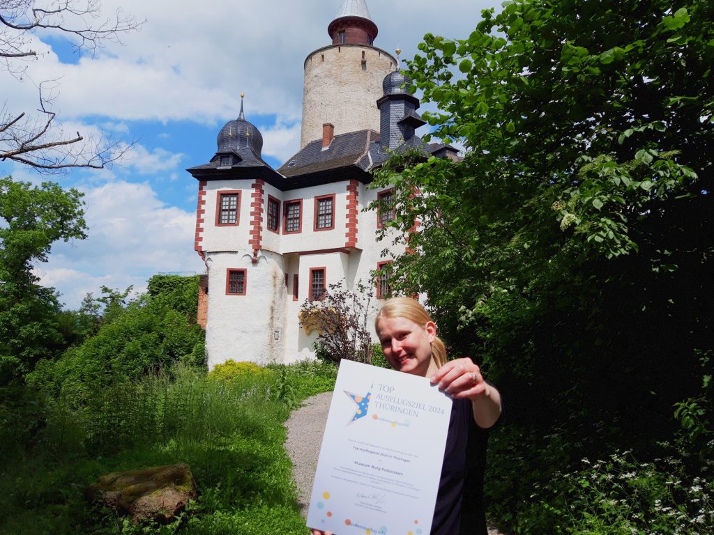 Eine blonde junge Frau hält eine Urkunde ins Bild, dahinter die Burg Posterstein und viele grüne Natur