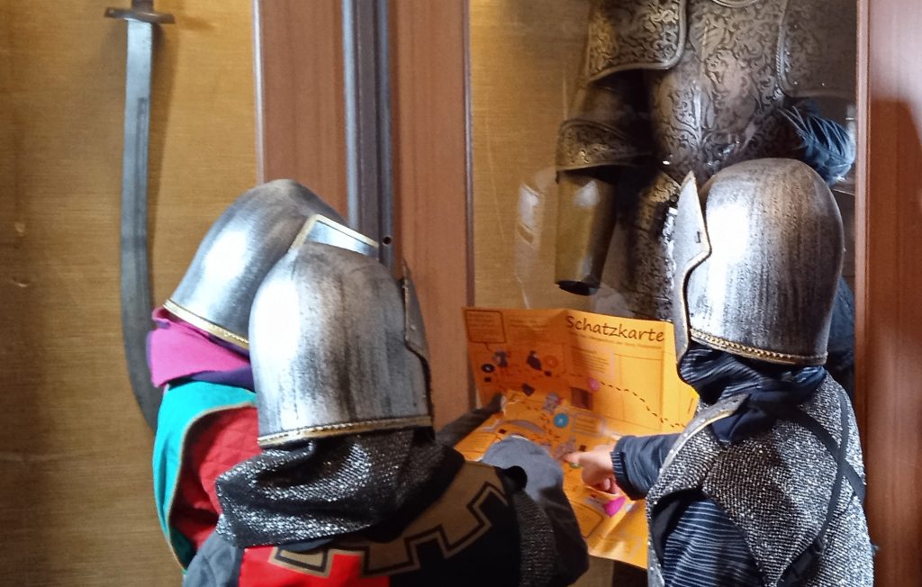 Drei Kinder in mittelalterlicher Verkleidung sehen sich die Schatzkarte durch die Kinderburg-Ausstellung auf Burg Posterstein an. Im Hintergrund eine Rüstung.