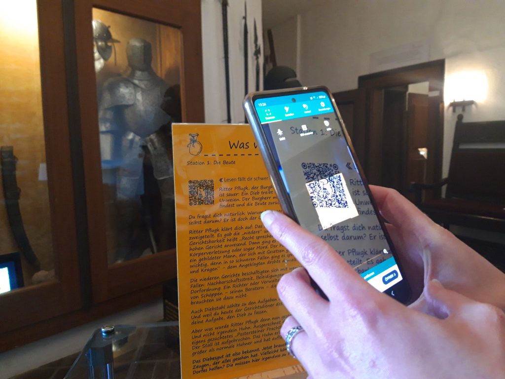 Im Vordergrund zwei Hände, die ein Smartphone halten und einen QR-Code scannen, im Hintergrund eine Ritterrüstung