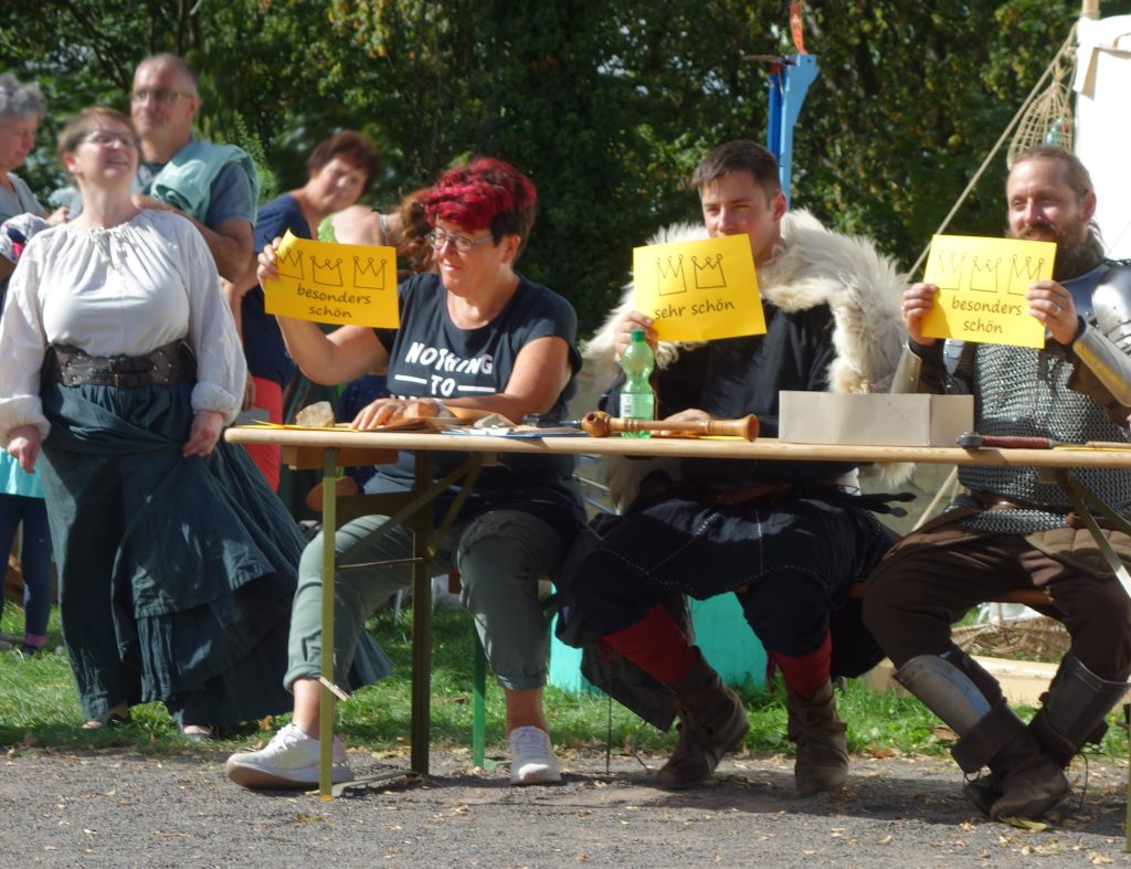 Eine Frau und zwei Männer in mittelalterlicher Gewandung halten gelbe Schilder hoch, auf denen "besonders schön" und "sehr schön" steht