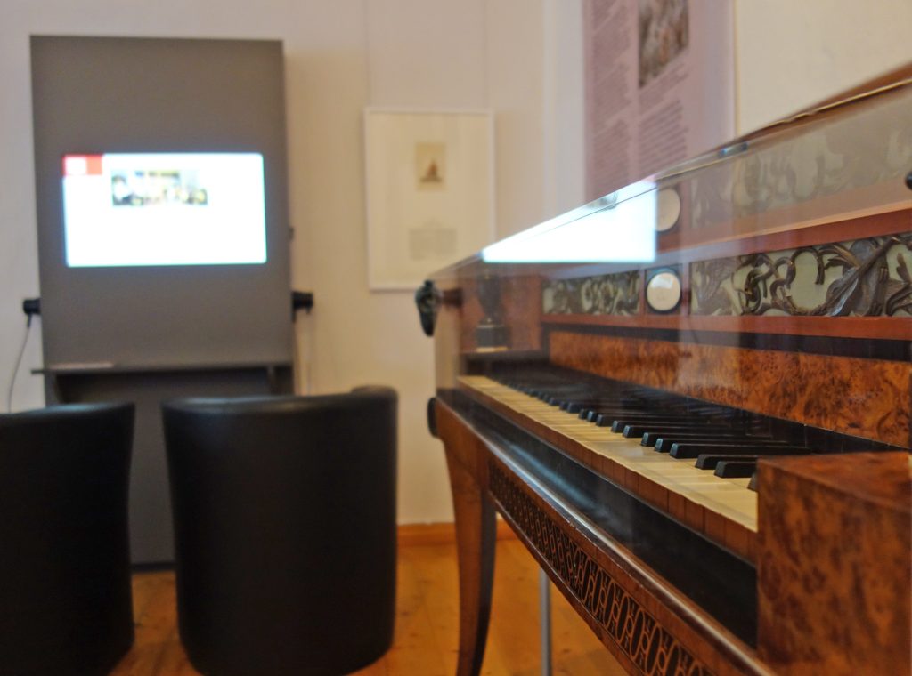 Historisches Klavier im Vordergrund, Medienstation mit zwei Sesseln davor im Hintergrund