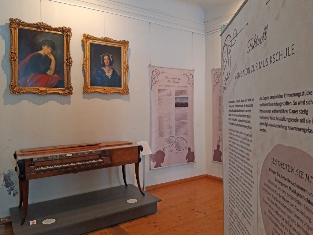 Rechts eine Ausstellungswand mit dem Ausstellungstitel, links ein historisches Klavier und zwei historische Gemälde im Goldrahmen